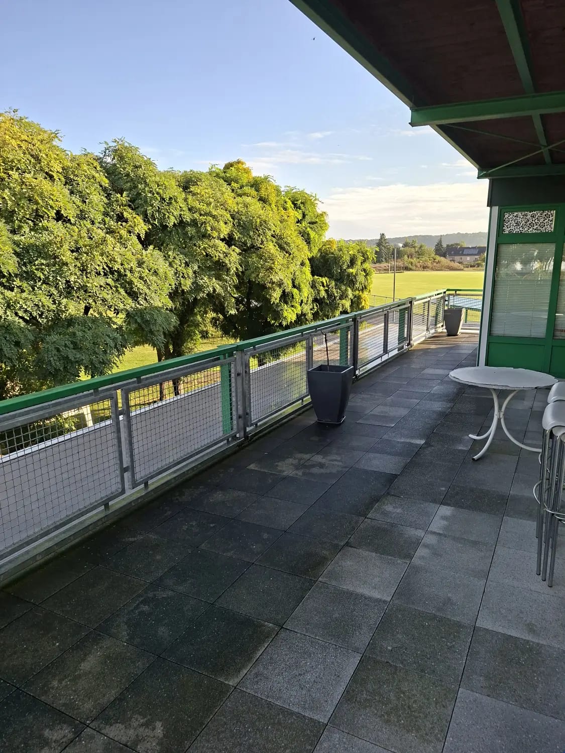 Terrasse im ersten Stock im Abendlicht, grünes Geländer, Bistrotisch und Blick über den Sportplatz