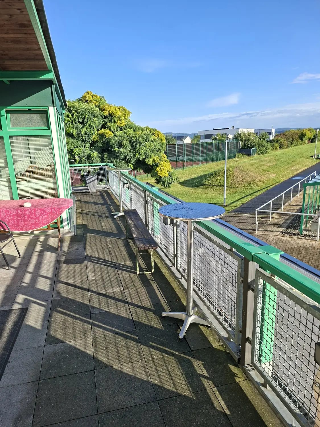 Terrasse mit Kübelbäumen entlang des grünen Geländers und Blick über das Sportgelände